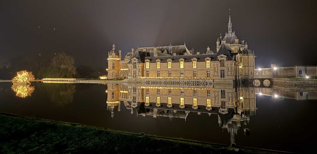 Le Château de Chantilly illuminé de nuit, avec ses décorations lumineuses se reflétant dans les douves à l'approche des fêtes de Noël.
