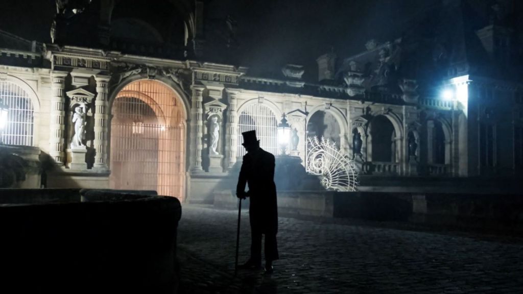 Halloween au Château de Chantilly : silhouette mystérieuse en haut-de-forme devant les Grandes Écuries illuminées, dans une atmosphère nocturne et brumeuse.